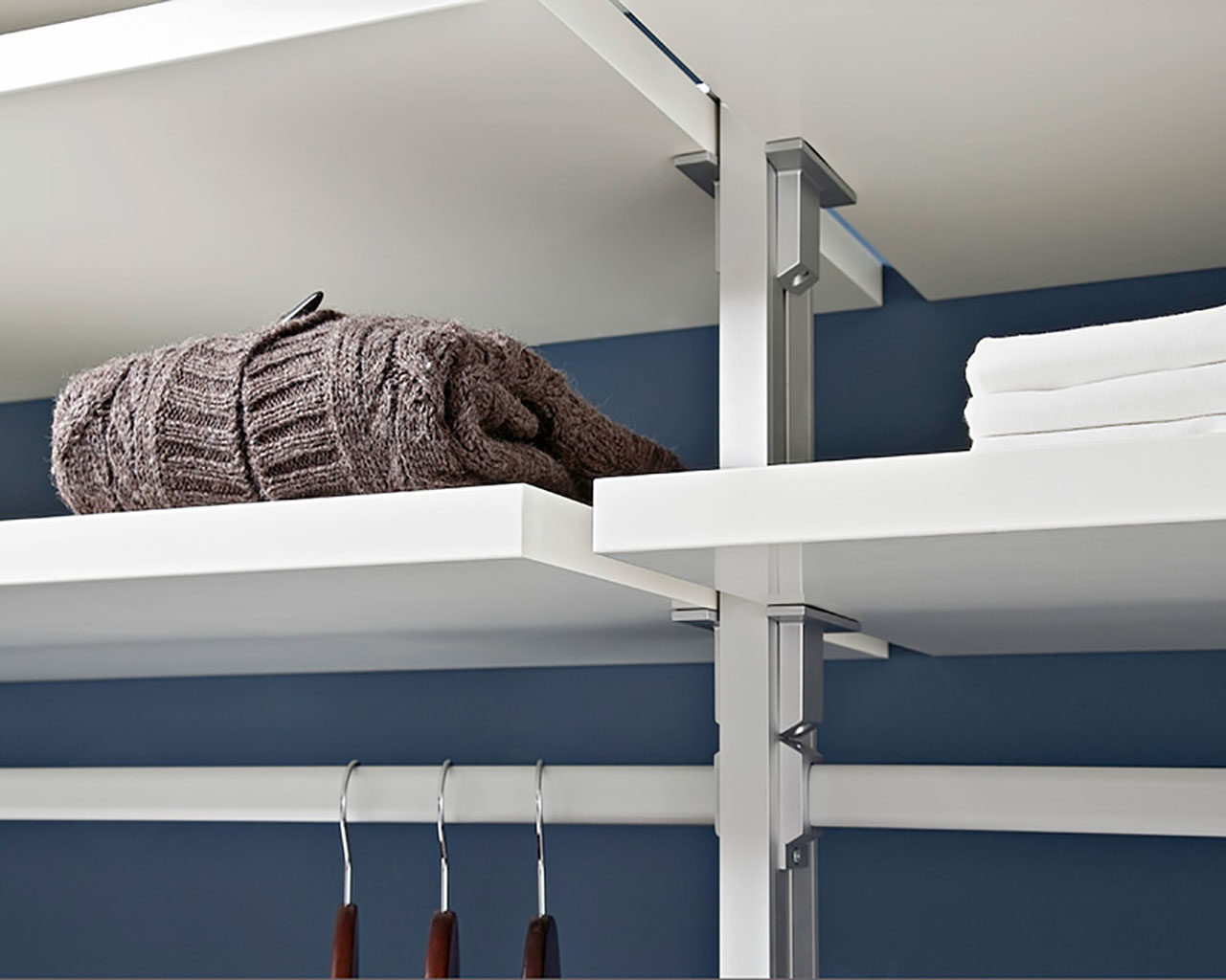 Ein Bildausschnitt vom Schranksystem Premium mit einem silbernen Profil an dem weiße Holzboeden sowie silberne Kleiderstangen mit Kleiderhaken befestigt sind. 