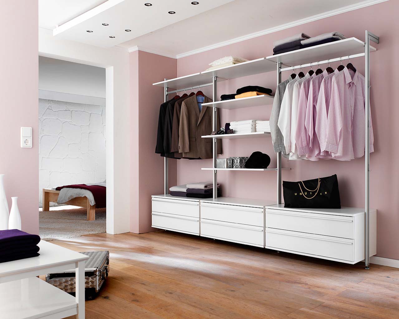 Ein stilvolles rosa Schlafzimmer mit einem eleganten weißen offenen Kleiderschrank ohne Türen mit vielen Schubladen, Regalen und Kleiderstangen, die den Raum perfekt ergänzen.