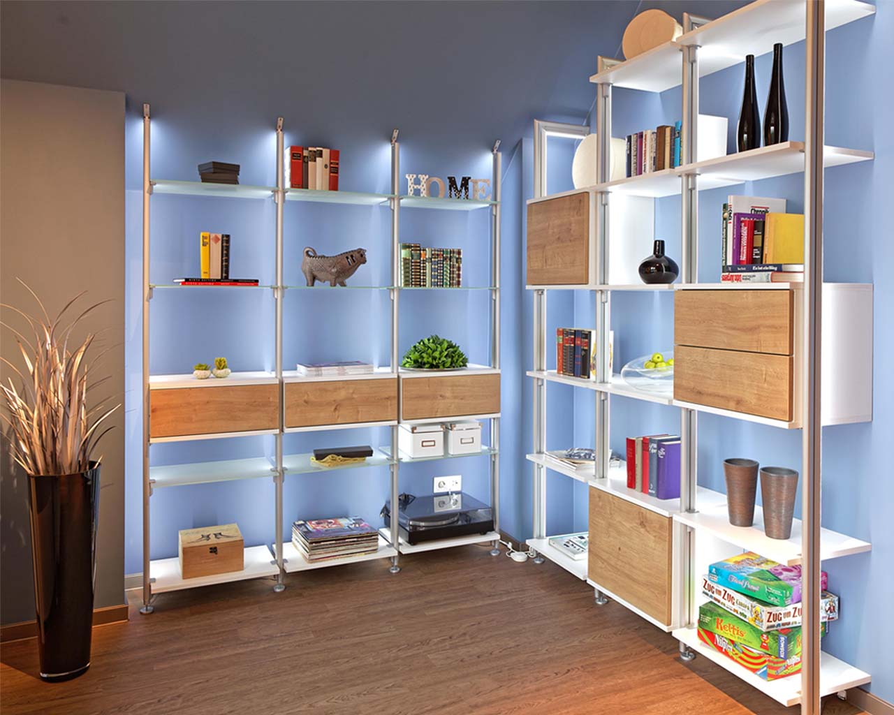 Ein gemütlicher Raum mit Regalen über zwei Wänden mit LED Beleuchtung und Holz- und Glasböden für Ablage von Büchern, Spielen, Plattenspieler mit Schallplatten, abwechselnd Schrankelemente mit Türen und Schubladen in Weiß, Frontfarbe Wildeiche, einer Bode